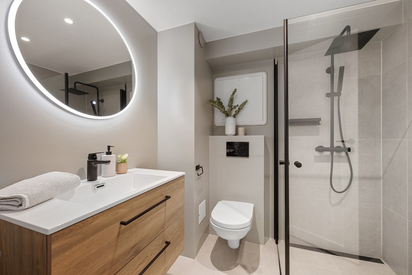 Stilrent og moderne bad med downlights og delikate kontraster. Innredning med store oppbevaringsskuffer, rundt speil med integrert belysning, sorte detaljer og dusj med slukrenne gir et gjennomført og eksklusivt uttrykk. Galleribilde