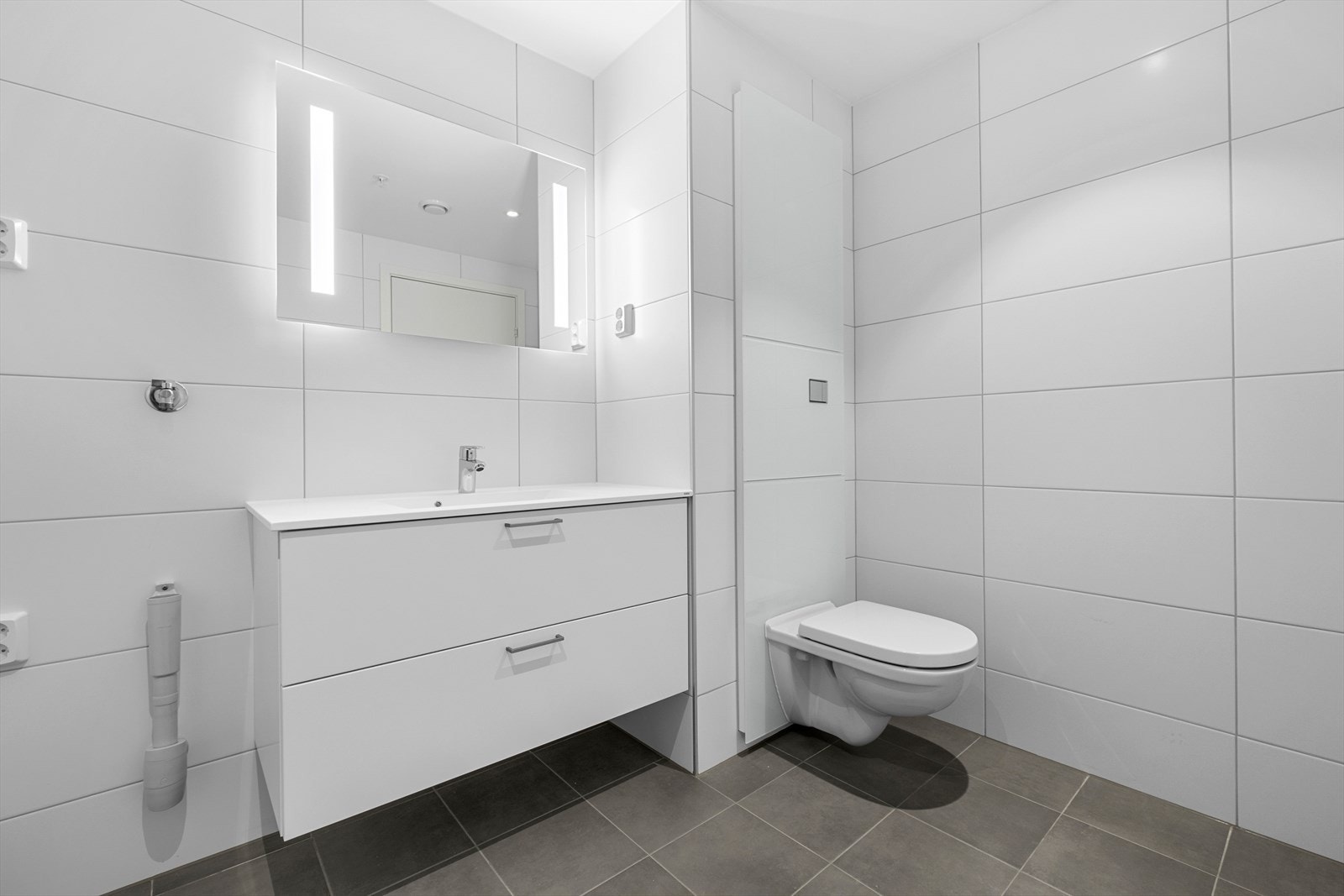 Baderommet er utstyrt med vegghengt toalett, behagelig gulvvarme, dusjhjørne med dusjgarnityr på vegg, samt opplegg for vaskemaskin. Galleribilde