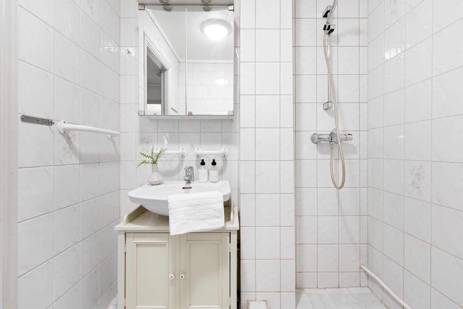 Badet er flislagt med lyse fliser på gulv og vegg og innredet med vegghengt vask med innredning under, speilskap, dusj og opplegg for vaskemaskin. Galleribilde