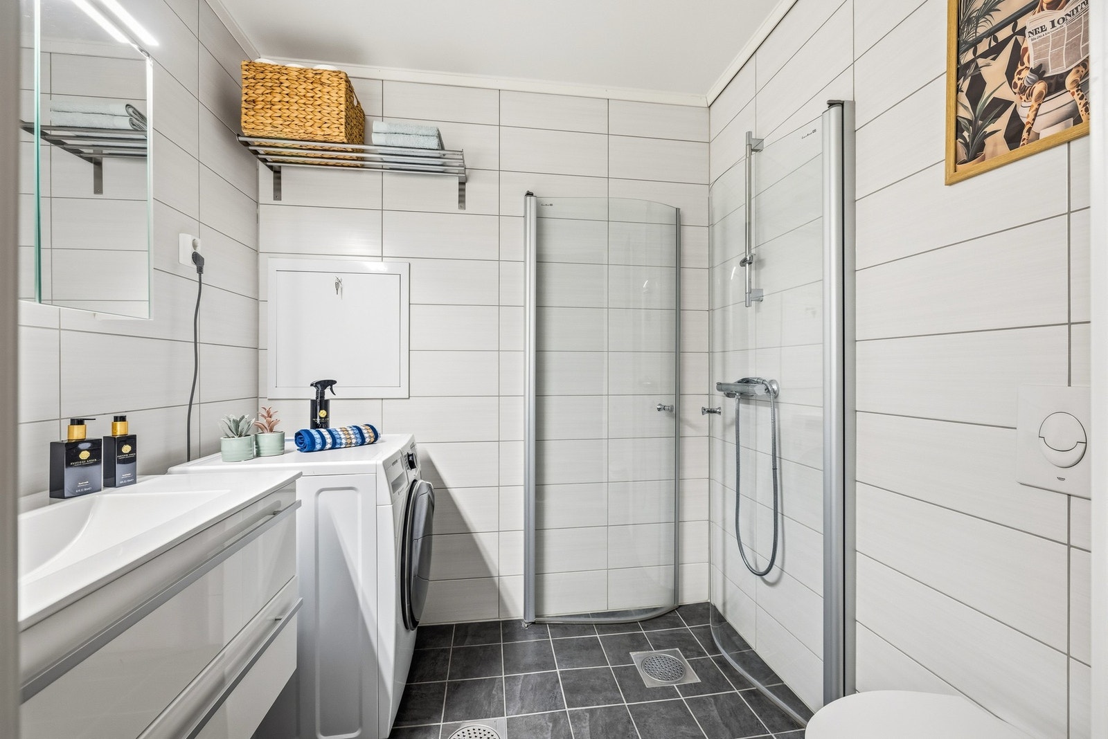 Baderommet har adkomst fra entréen og har fliser på gulv og vegger med elektrisk gulvvarme. Baderommet er utstyrt med vegghengt toalett, dusjhjørne med innfellbare glassdører og dusjarmatur, samt opplegg for vaskemaskin og tørketrommel. Galleribilde