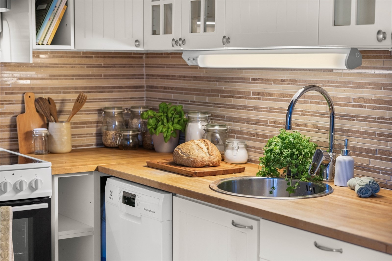 Innredning med profilerte fronter og heltre benkeplate med nedfelt vaskekum, samt overskap og fliser på vegg over benkeplaten. Galleribilde