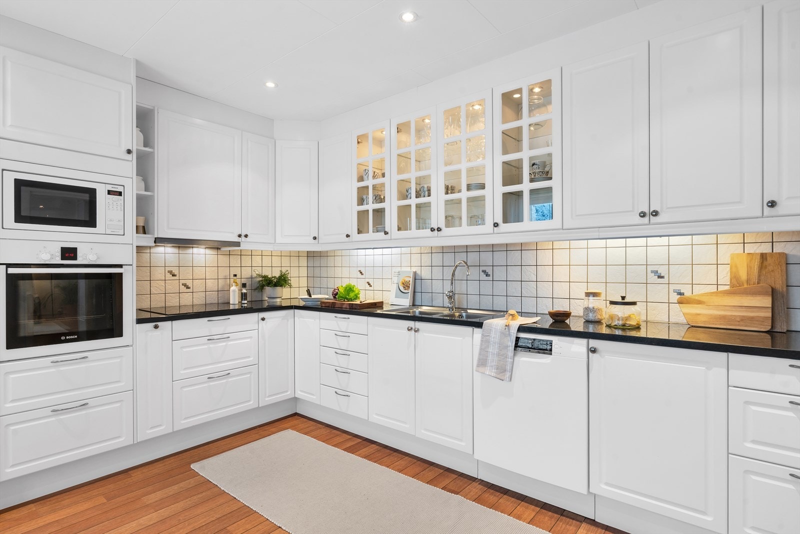 Innbydende kjøkken i hvit utførelse med profilerte fronter, fliser på vegg over benkeplate, samt lekker benkeplate av stein. Galleribilde