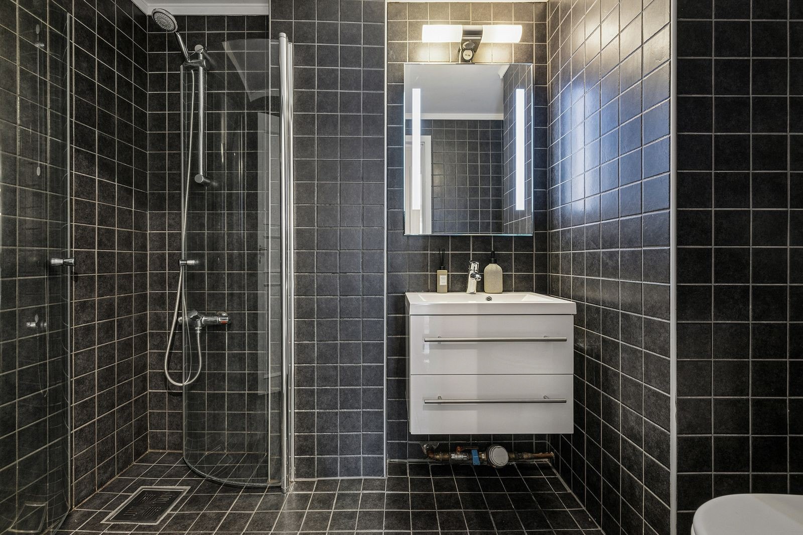 Rommet er innredet med gulvstående toalett, dusjhjørne med dusjvegger og en servantinnredning med skuffer. Over servanten er det montert speil med integrert belysning. Galleribilde