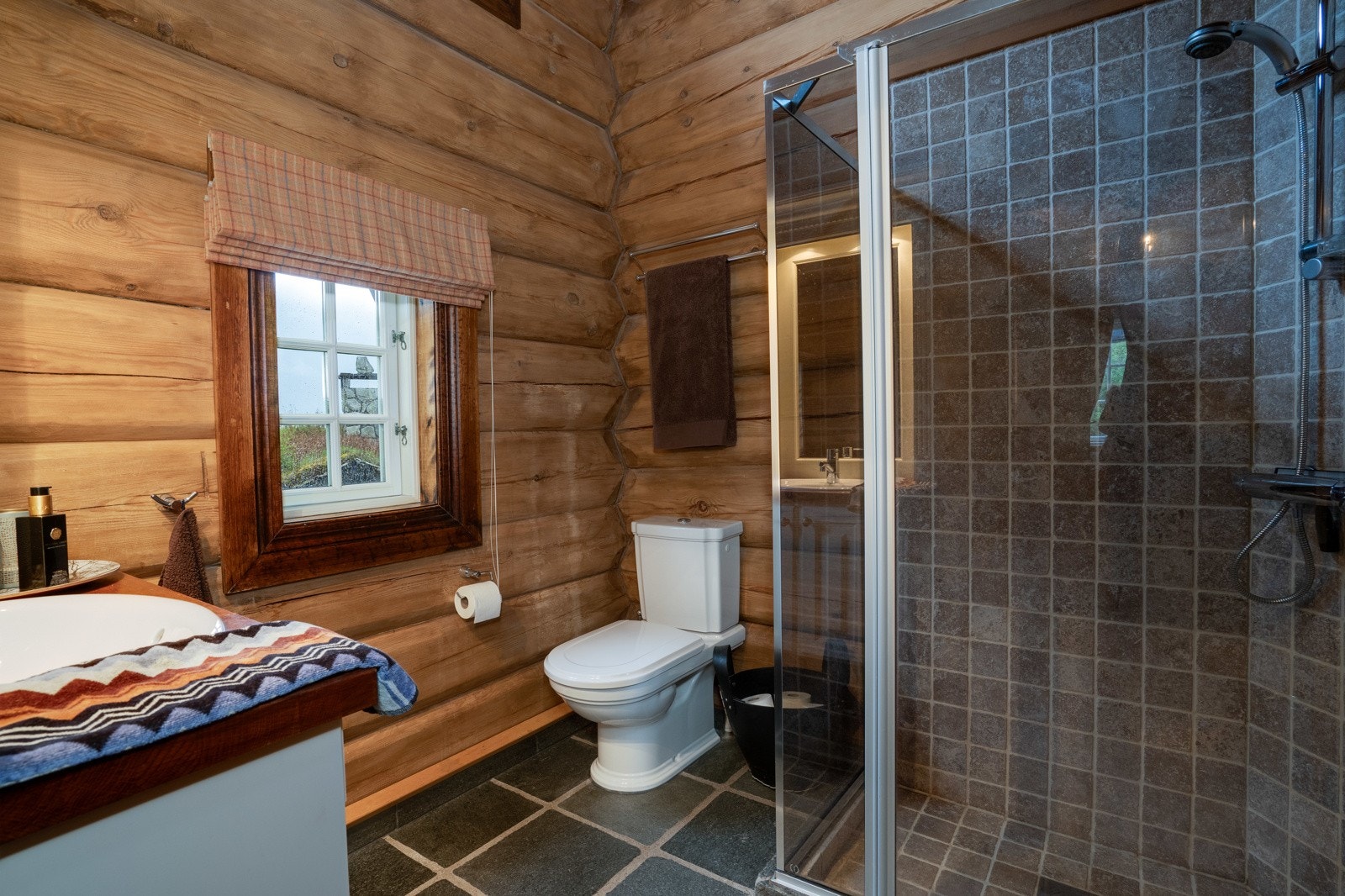 Hytta inneholder to delikate baderom, badstue og et praktisk vaskerom. I tillegg er det et bad i annekset. Badene i hytta er holdt i samme stil med store skiferfliser på gulv, tømmervegger, samt delikate, sandfargede fliser i dusjsonen. Galleribilde