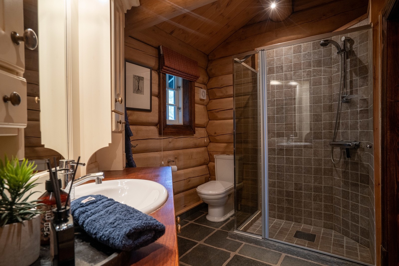 Bad 2 med store skiferfliser på gulv og flotte tømmervegger, samt delikate, sandfargede fliser i dusjsonen. Galleribilde