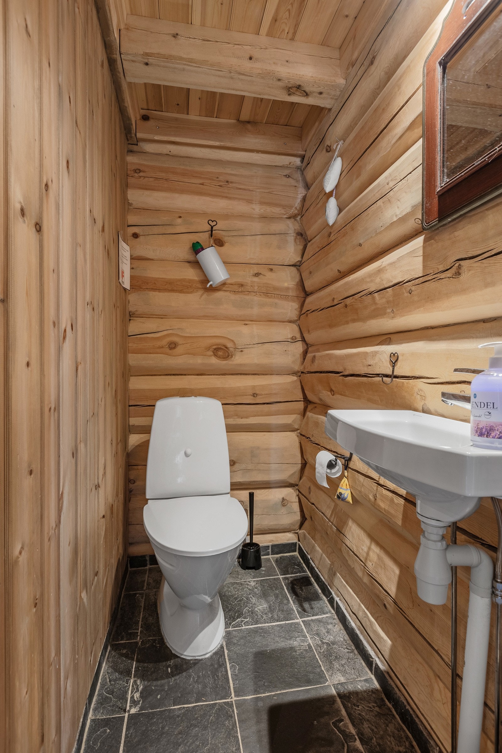 Toalettrommet har også fliser på gulv, og en kombinasjon av tømmer og panel på veggene og i taket. Rommet inneholder et gulvstående toalett og en enkel servant. Galleribilde