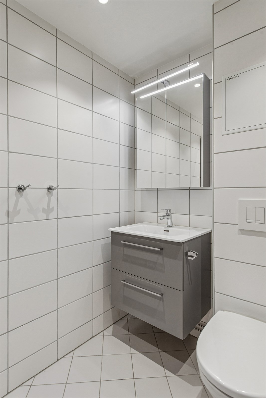 Rommet er innredet med en baderomsinnredning med servant og ettgreps blandebatteri, vegghengt toalett og et dusjhjørne med glassdører. Det er også opplegg for vaskemaskin. Galleribilde