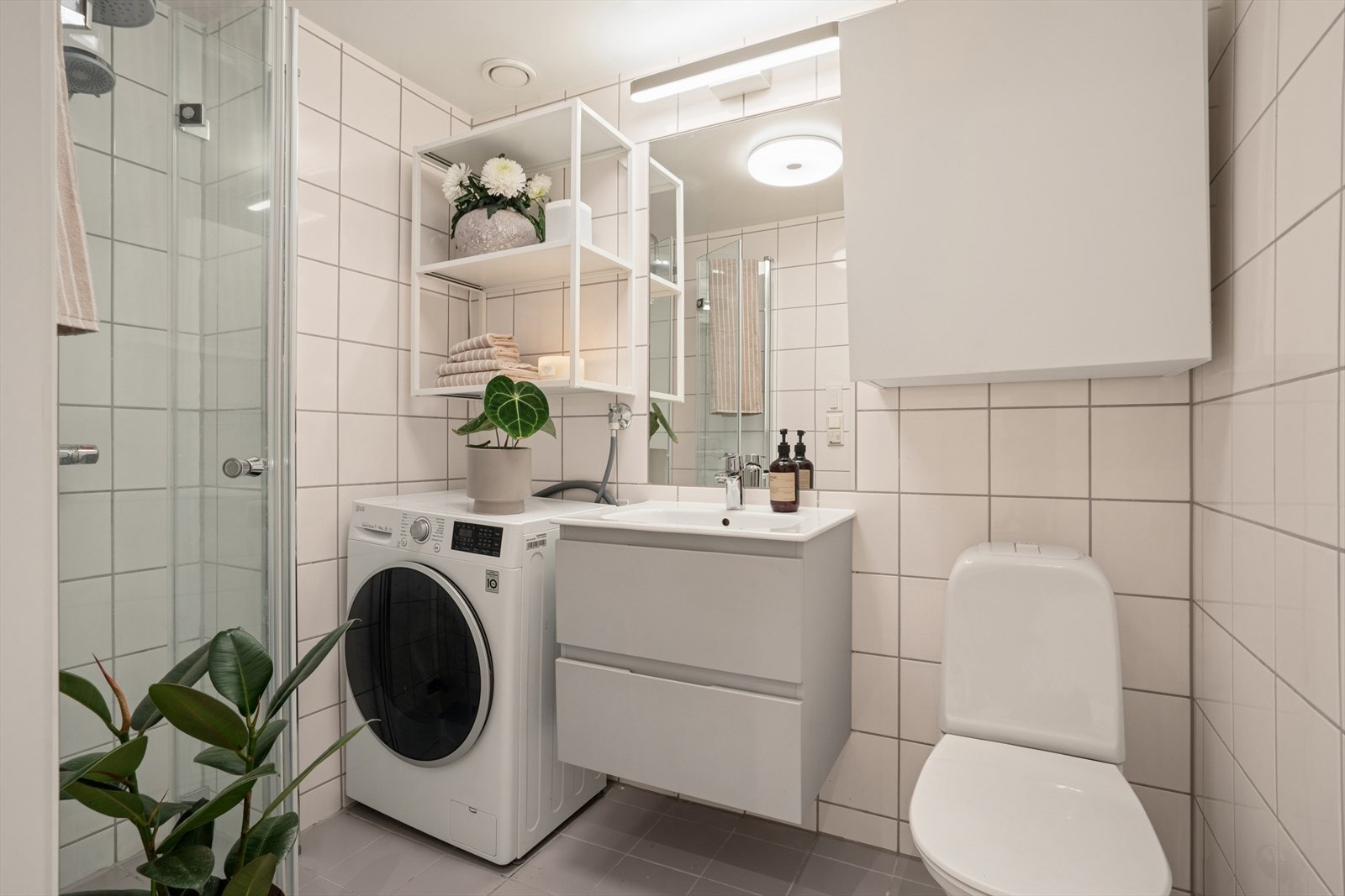 Pent flislagt baderom med toalett, servant med underskap og overspeil, dusj med innsvingbare dører og opplegg for vaskemaskin. Galleribilde