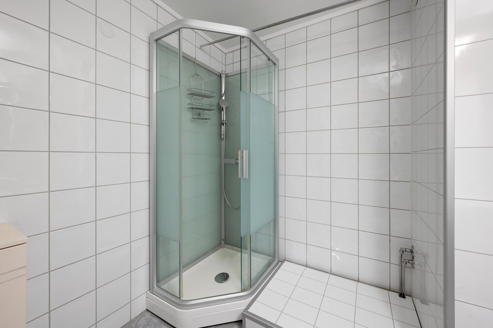 Dusjkabinett plassert i hjørnet med hvite vegg- og gulvfliser. Galleribilde