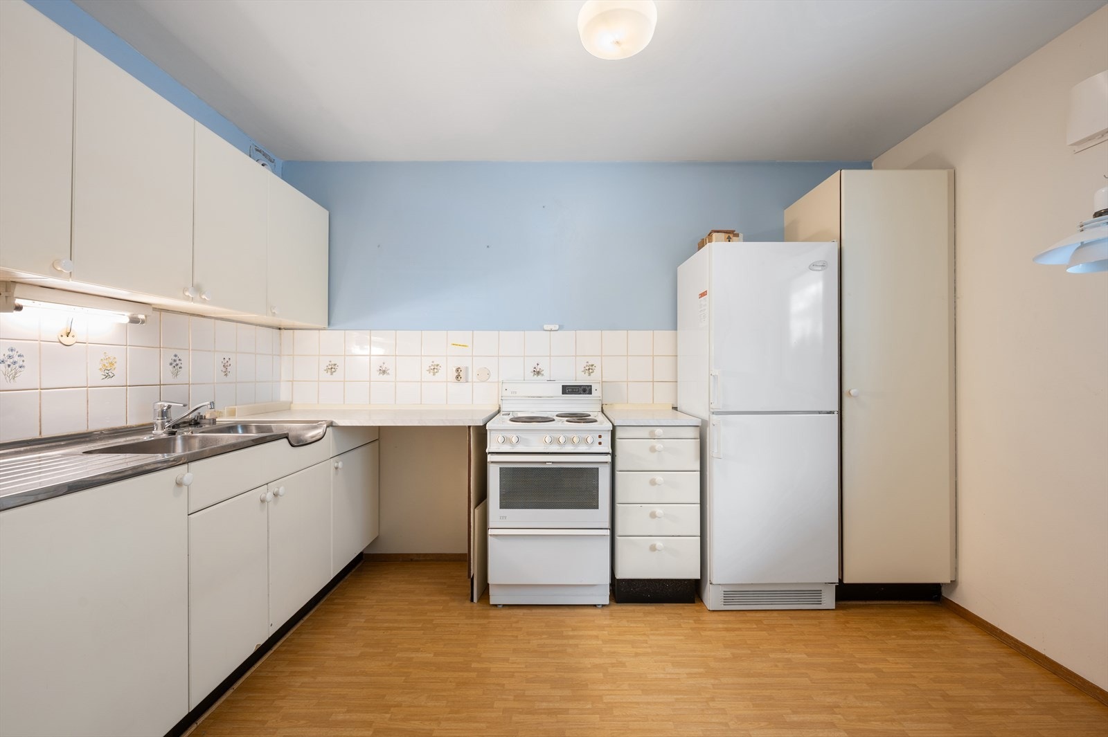 Kjøkken med eldre innredning, frittstående hvitevarer og laminatgulv. Galleribilde