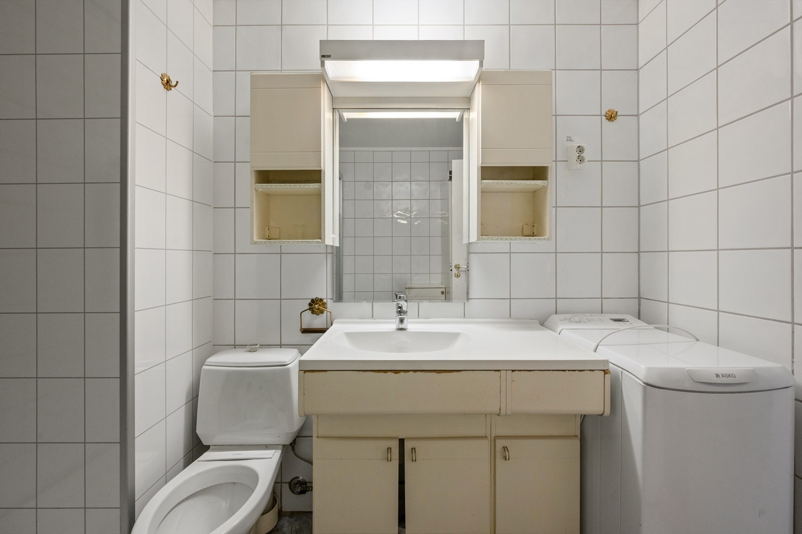 Bad med veggfliser, toalett, servantskap og opplegg til vaskemaskin - innredningen og badet bærer preg av slitasje og modernisering er nødvendig. Galleribilde