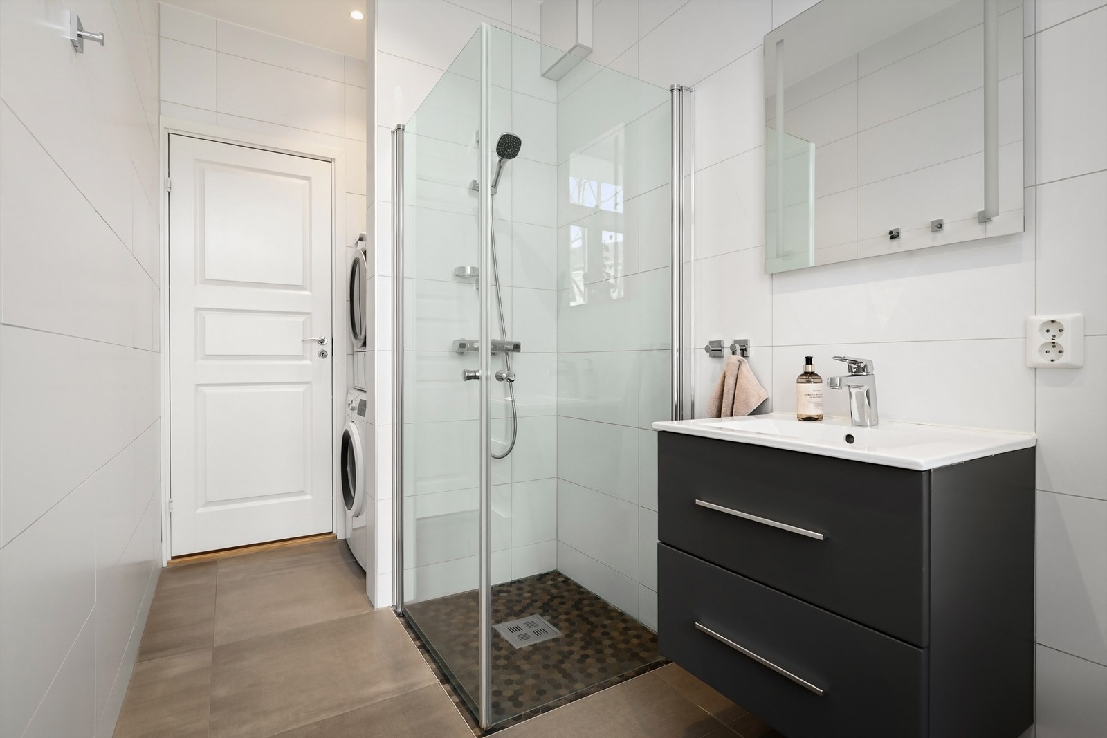 Videre er badet innredet med dusjhjørne med innfellbare glassdører og vegghengt toalett. Opplegg og plass for vaskemaskin og tørketrommel i praktisk nisje Galleribilde