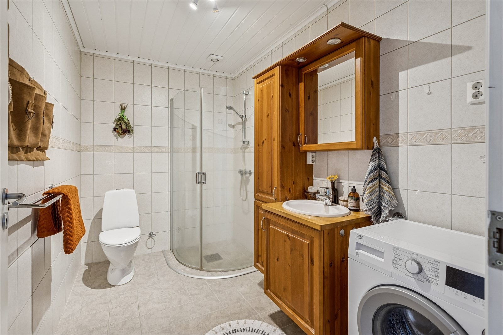 Flislagt bad med servantskap, høyskap, gulvmontert WC og et dusjhjørne med glassdører. Galleribilde