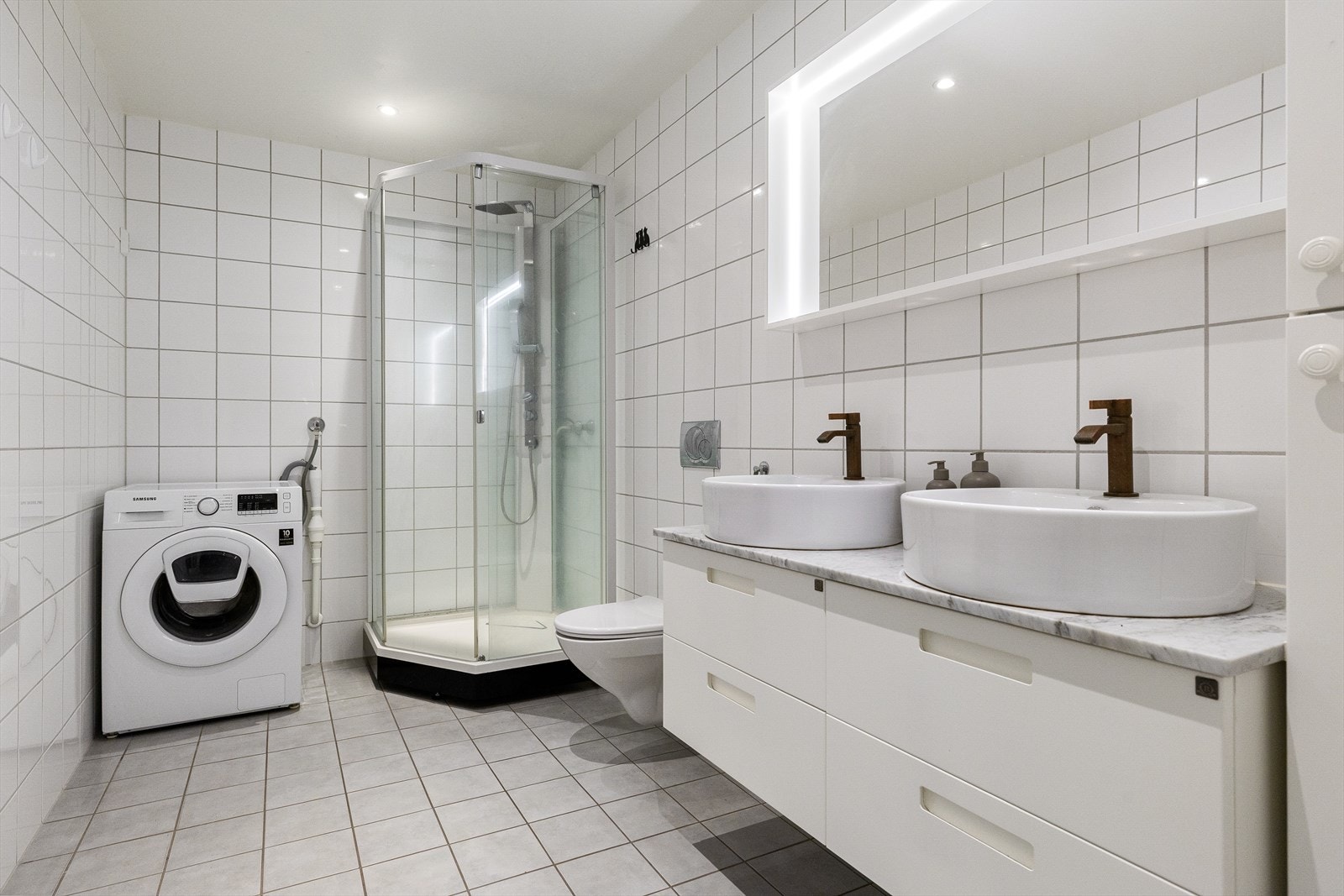 Leilighetens bad er flislagt med varme i gulvet. Badet tilbyr god plass og er utstyrt med to ovenpåliggende servanter montert på innredning, veggmontert toalett, og et praktisk dusjkabinett. Galleribilde