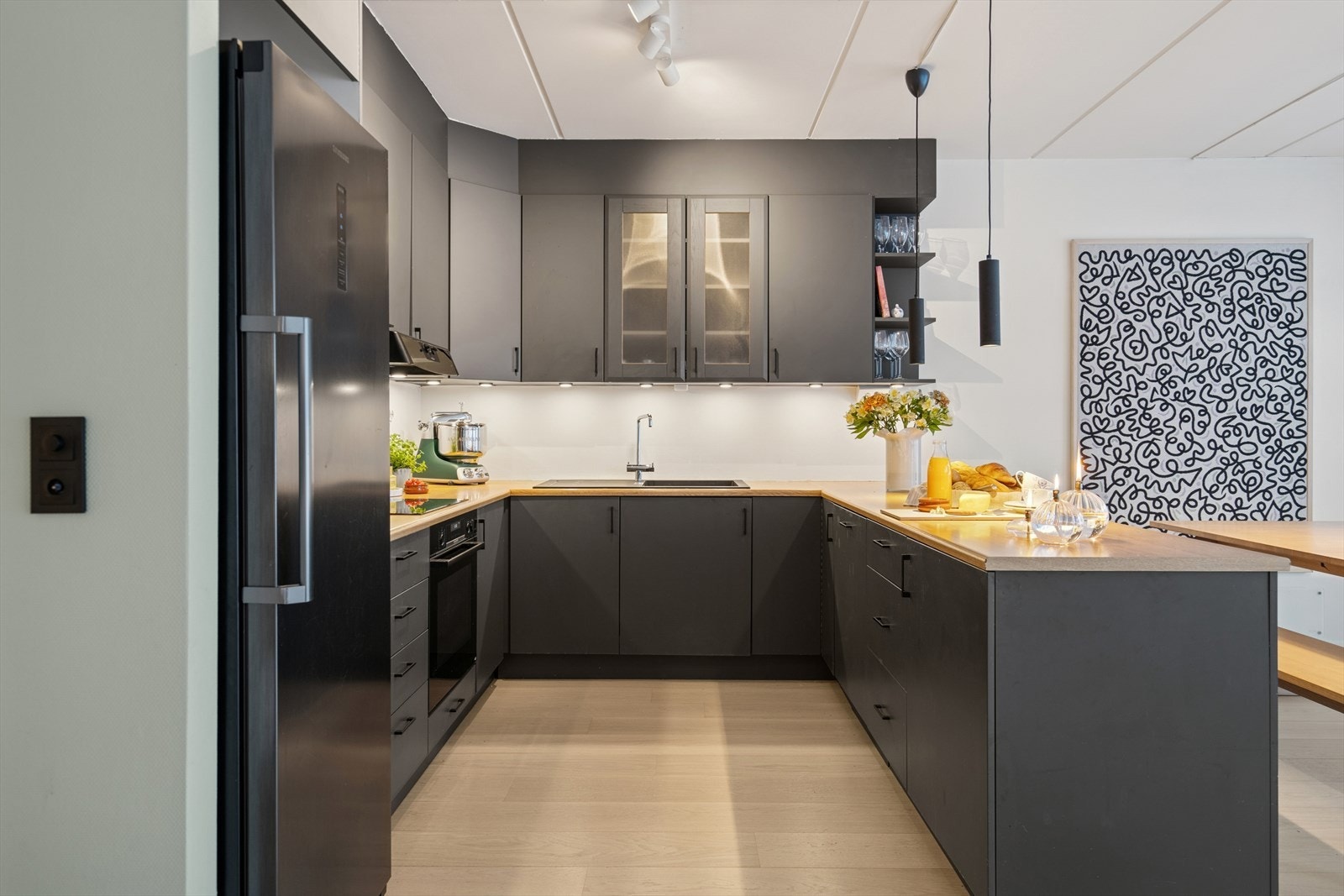 Kjøkkenet har en moderne, mørk innredning med glatte fronter, samt et stilfullt overskap med glassfronter. Her får du god plass til matlagingen med mye skap- og benkeplass. Galleribilde