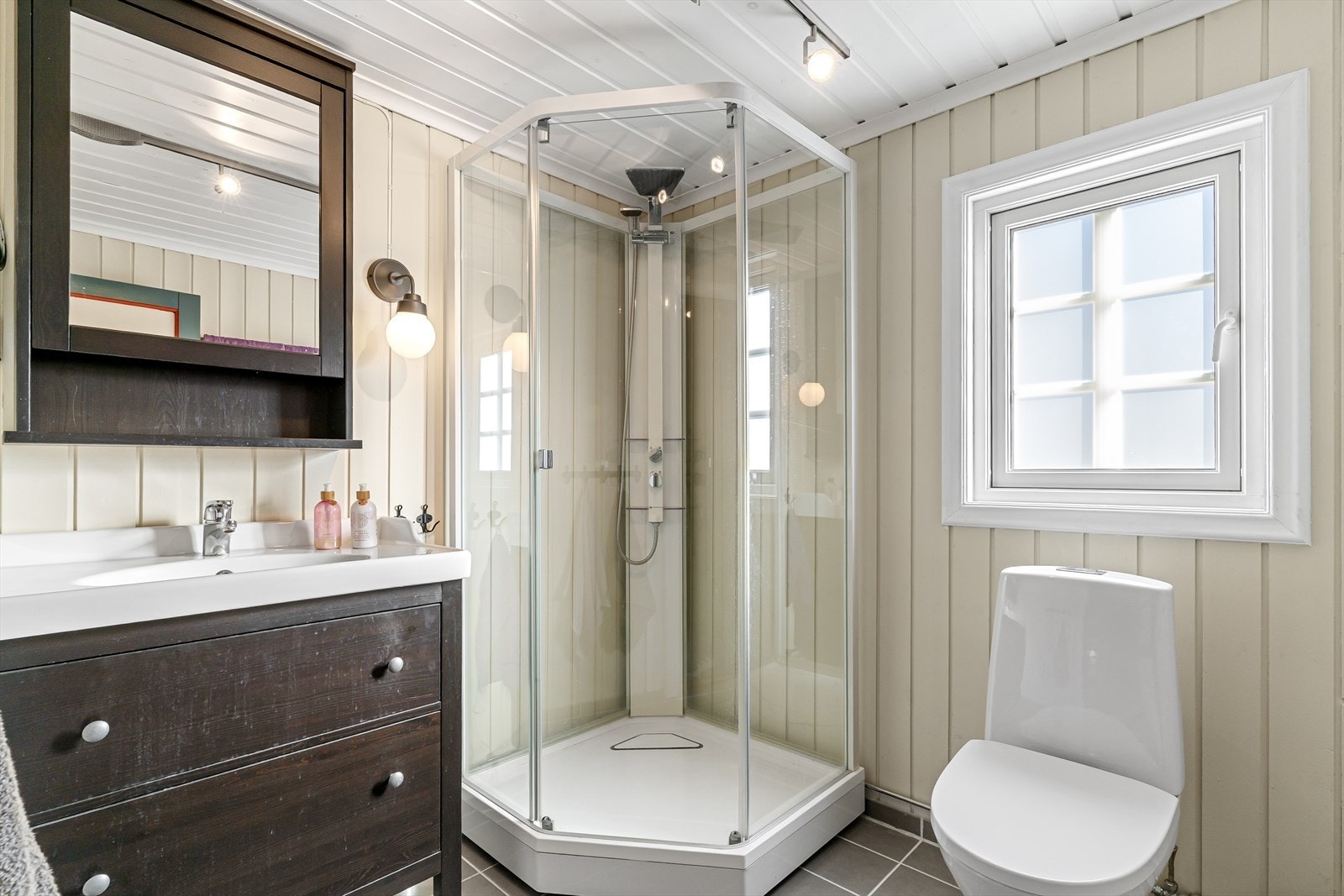 Badet fremstår med tidsriktige fargevalg og har flislagt gulv med varmekabler for ekstra komfort. Rommet har innredning med nedfelt servant, toalett og dusjkabinett. Galleribilde