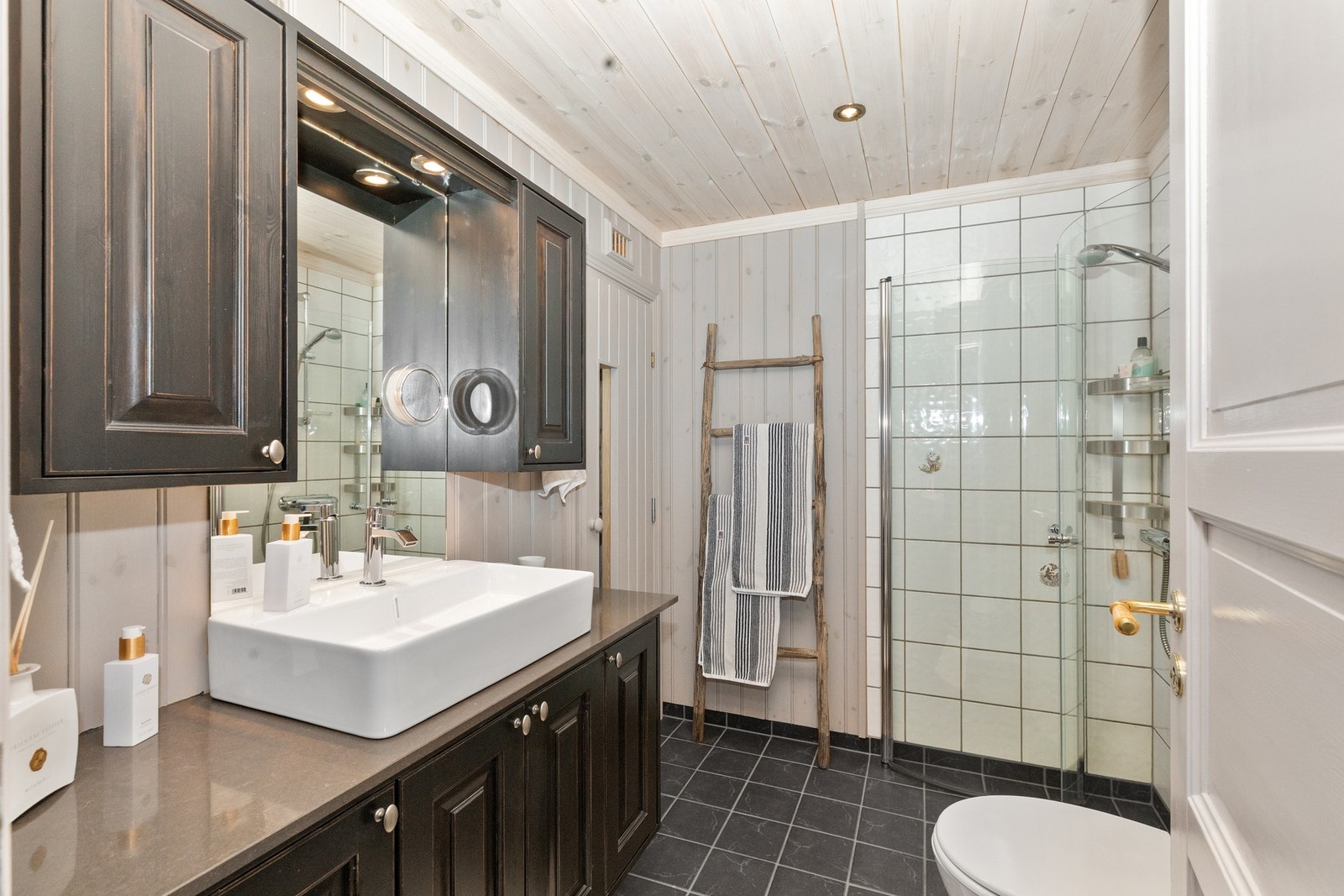 Flis på vegg i dusjhjørnet, panel for øvrig. Badet er møblert med servant med underskap, speilskap, dushjørne med dusjdører og gulvmontert toalett Galleribilde