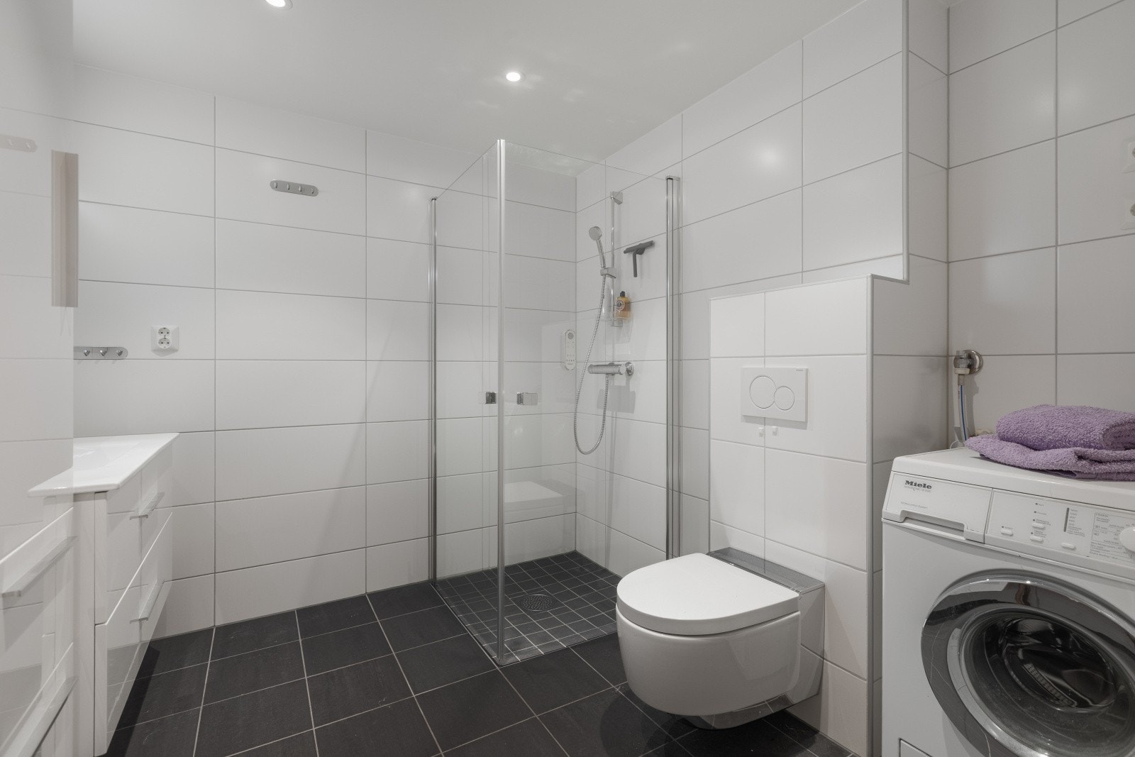 Badet er utstyrt med vegghengt toalett, servant, dusj og opplegg for vaskemaskin. Det er fliser på veggen og det er satt inn downlights i taket. Galleribilde