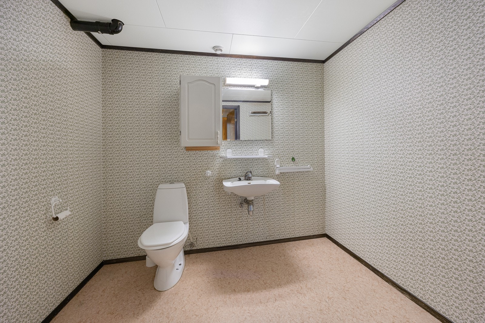 Wc-rommet har belegg på gulvet og tapetserte vegger. Videre er det enkel servant, veggskap, speil med belysning og gulvmontert toalett. Galleribilde