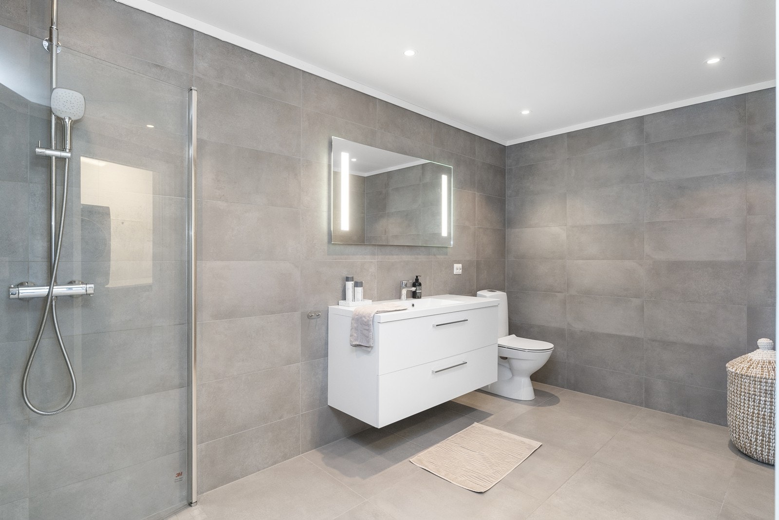 Badet har nøytral og tidløs baderomsinnredning med servant og vegghengt speil med belysning, wc og dushjørne. Galleribilde