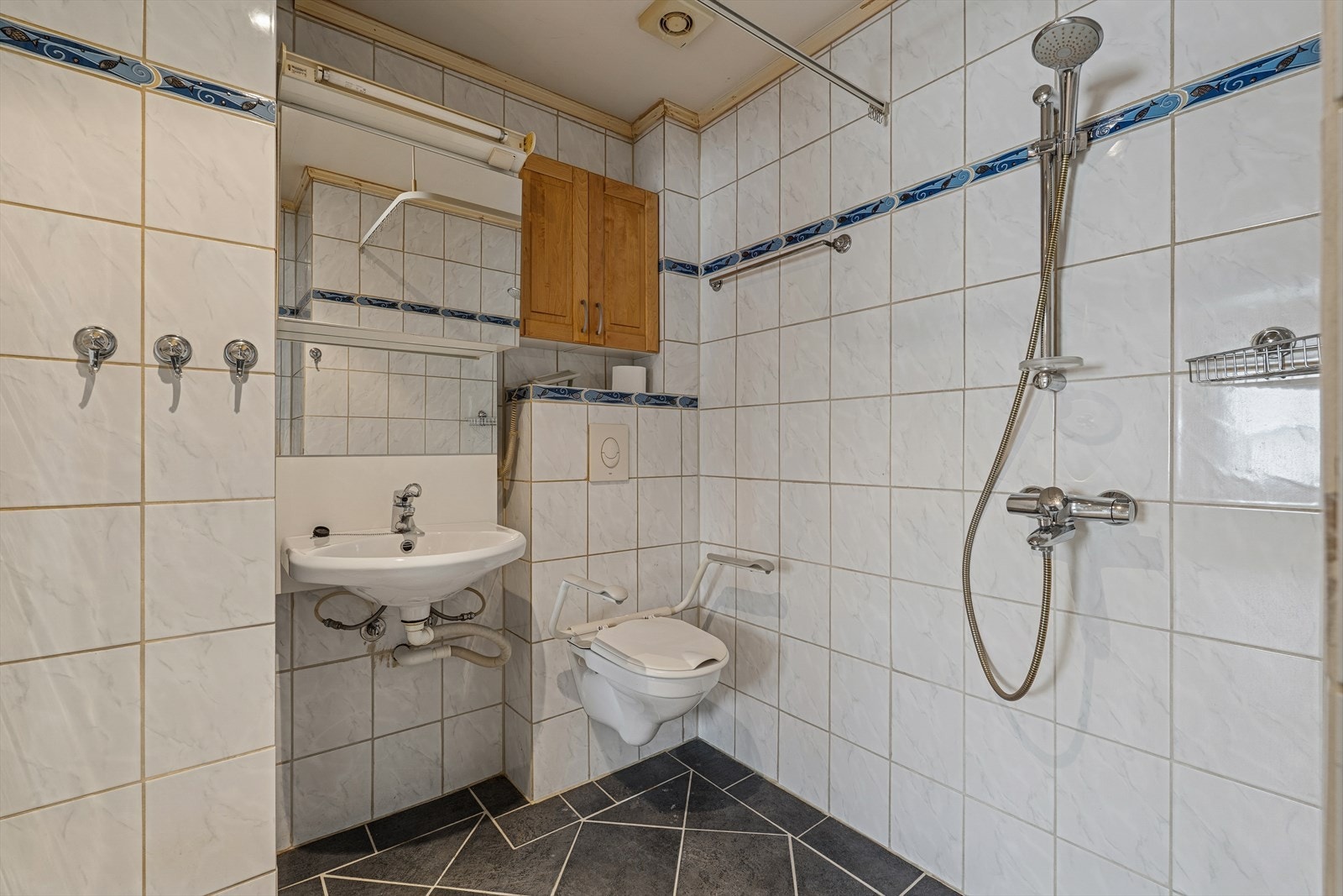 Baderom med flislagte overflater og dusj med forheng. Rommet er utstyrt med vegghengt toalett og servantløsning med elektrisk hev- og senkefunksjon, samt oppbevaring i overskap. Et funksjonelt baderom som gir et godt utgangspunkt for egne tilpasninger Galleribilde