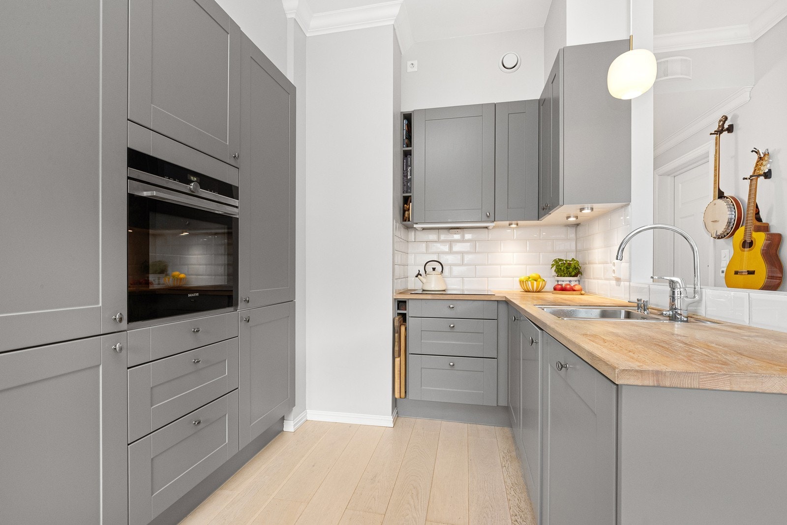 HTH kjøkkeninnredning med profilerte, grå fronter, heltre benkeplate og fliser på vegg over benk. Integrerte hvitevarer som oppvaskmaskin, komfyr, platetopp og kombinert kjøl-/fryseskap. Galleribilde