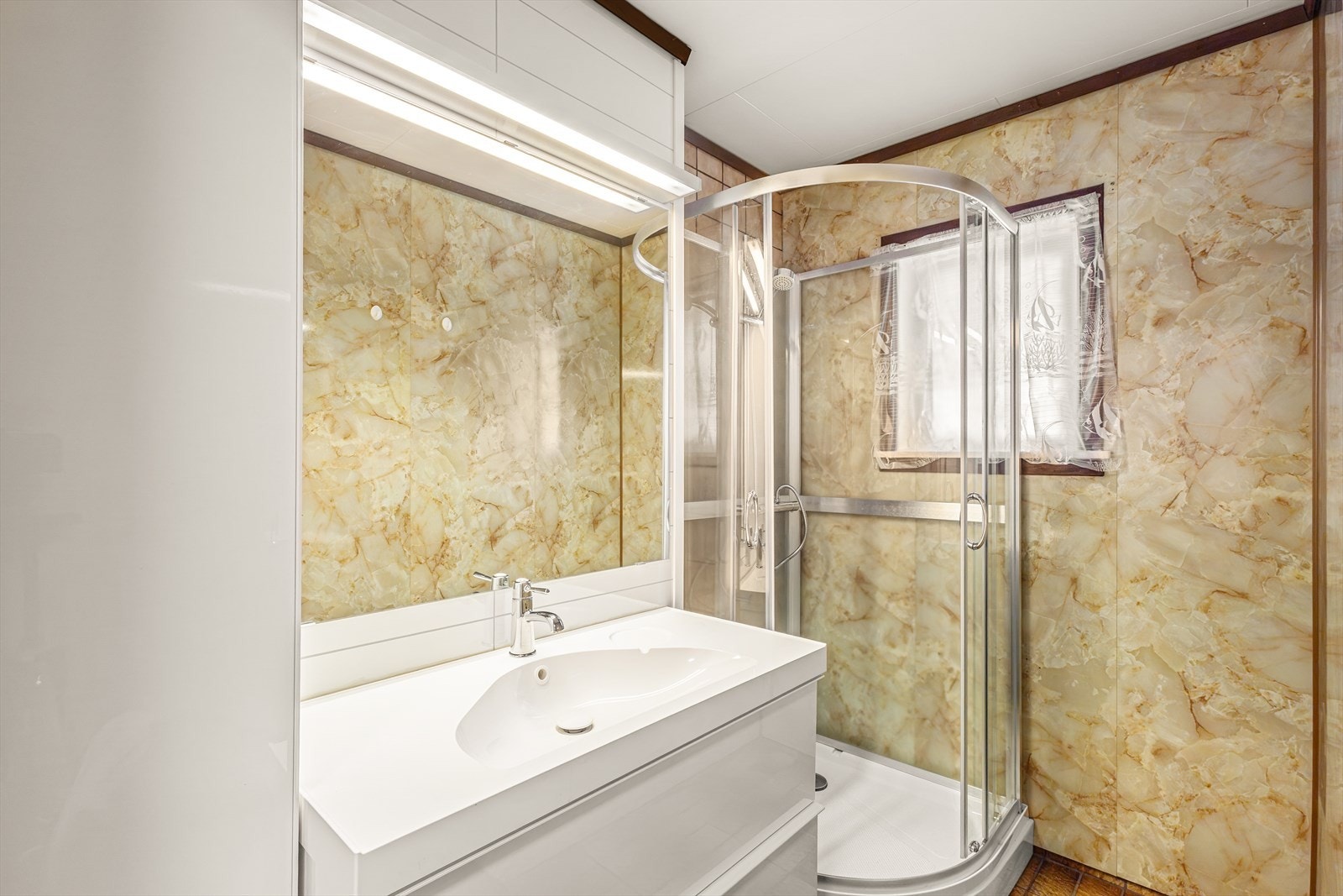 Badet er fra byggeår og har fliser på gulv og våtromsplater på vegg, med eldre sluk og naturlig ventilasjon Galleribilde