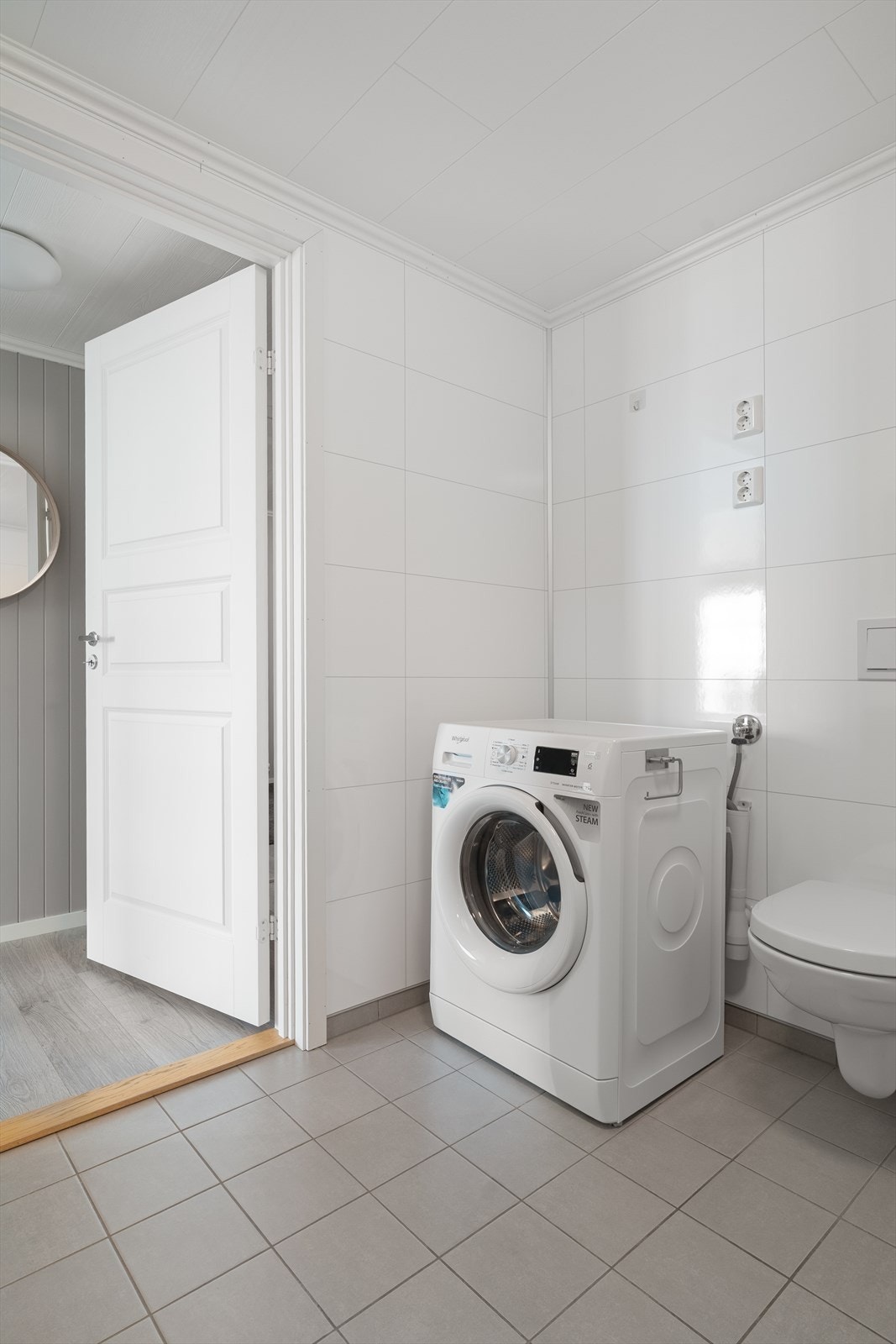 Badet er utstyrt med innredning med servant, dusjhjørne med glassdører og dusjgarnityr på vegg, vegghengt toalett og opplegg for vaskemaskin Galleribilde