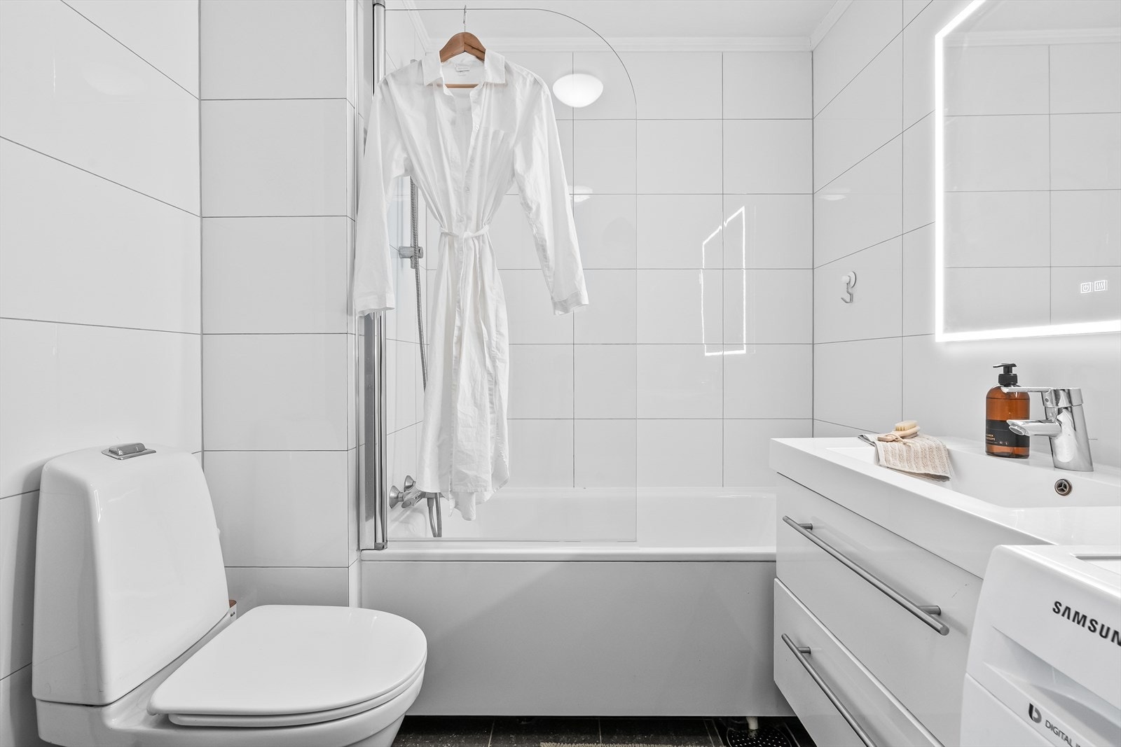 Badromsinnredning med skuffer, heldekkende servant og speil med lysarmatur, klosett og badekar med dusjutstyr og svingbar glassvegg Galleribilde