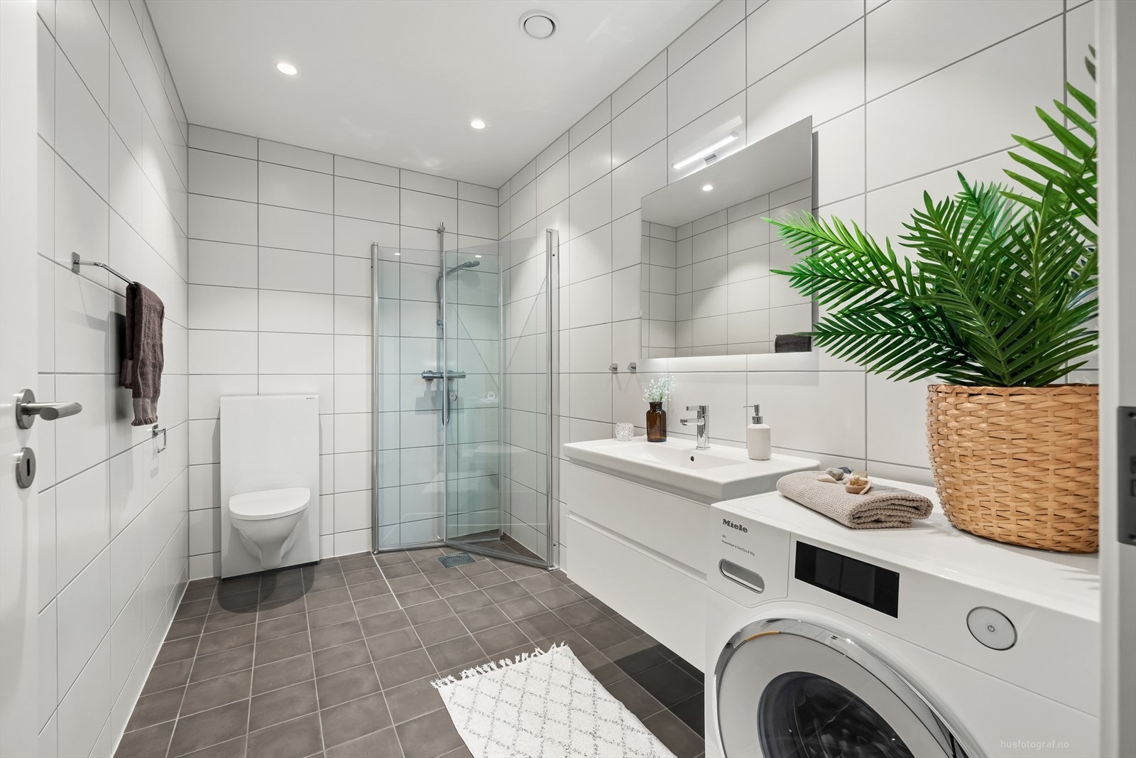 Det er dusj i hjørnet med innfellbare dører, vegghengt toalett og servantinnredning med skuffer og speil med overlys. Galleribilde