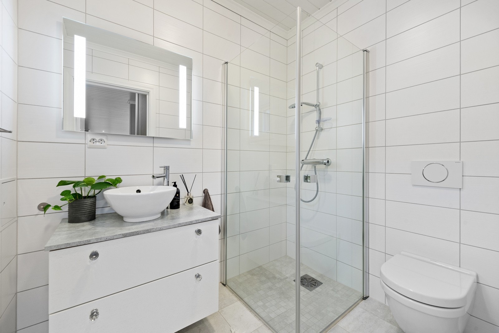 Baderom 2 i 1.etg ligger i gangen mellom stue og soverom 2. Et tidløst og fint baderom med fliser på vegger og gulv, vegghengt toalett, dusj med glassdører, innredning med skuffeløsning og speil med lys. Galleribilde