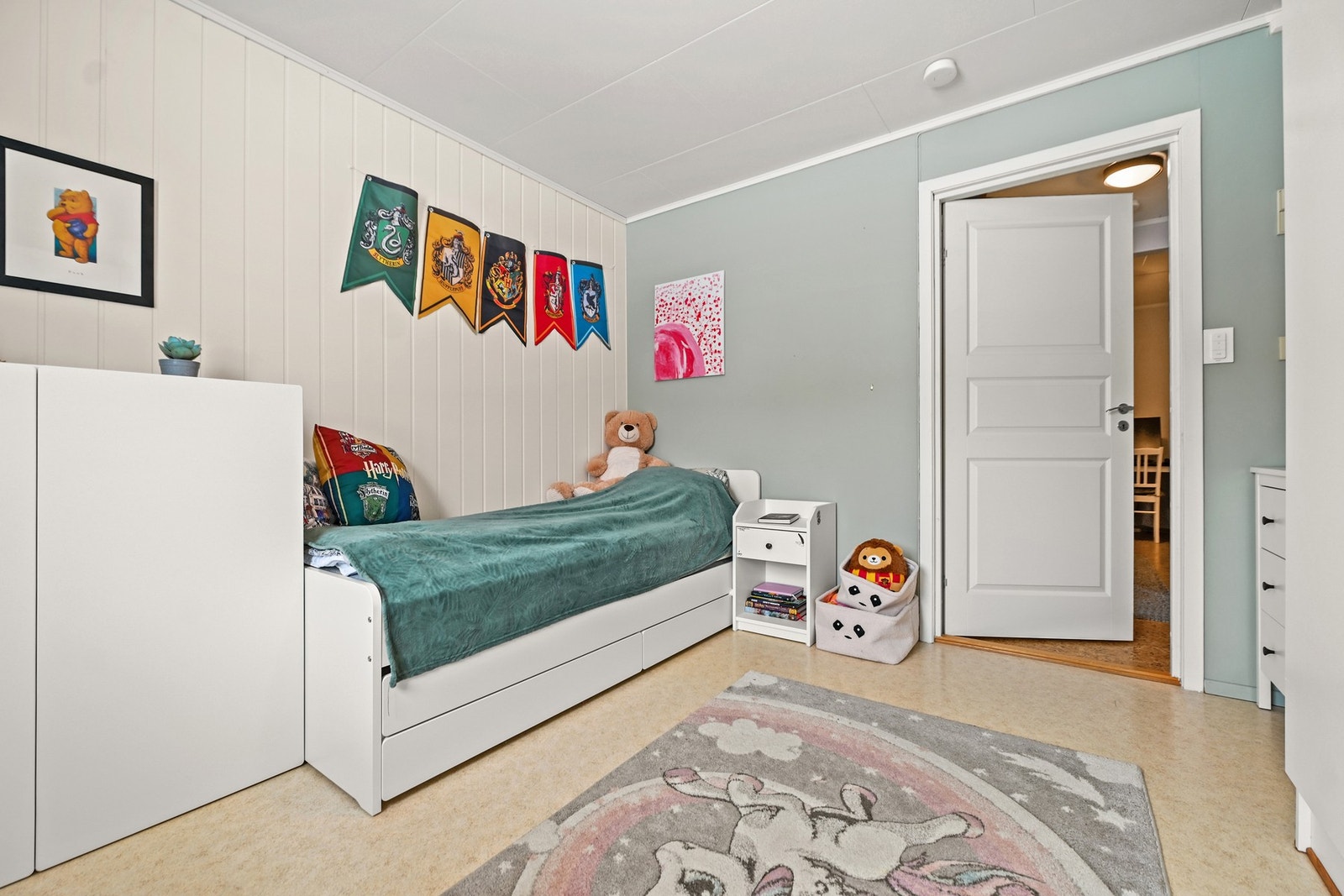 Barnerom med lyse vegger, dekorasjoner og en seng. Dør tilknyttet rommet åpner mot et annet rom. Galleribilde