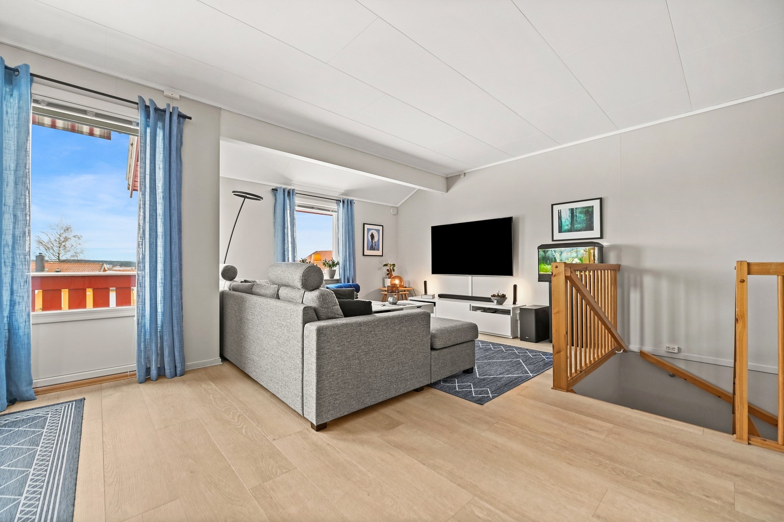 Lys stue med store vinduer, lyse vegger og parkettgulv. Veggmontert TV og åpen trapp til underetasjen. Galleribilde