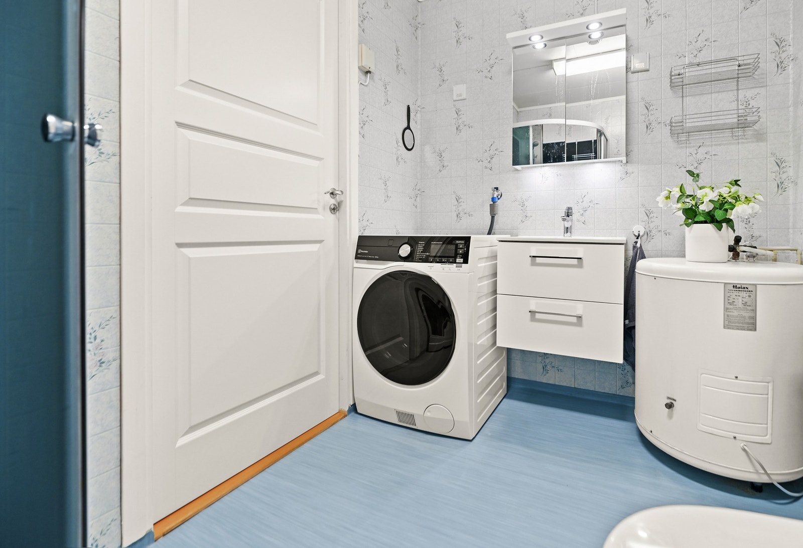 Baderom med vaskemaskin, varmtvannsbereder, servant med underskap og speilskap. Vegger med lys tapet og blått gulvbelegg. Galleribilde