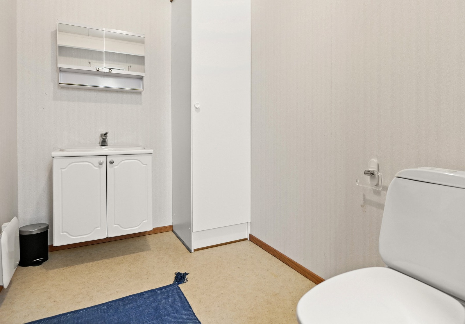 Badet har lyse vegger, servant med underskap, speilskap, toalett og et høyt skap for oppbevaring. Galleribilde