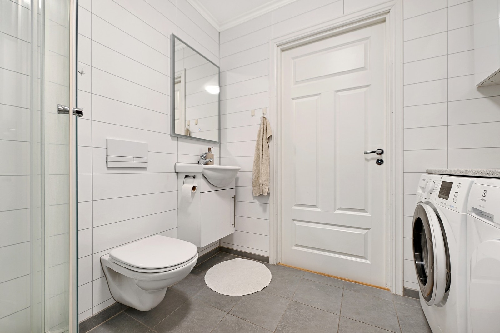 Bad med dusjkabinett, vegghengt toalett, vaskemaskin og servant. Lyse fliser på veggene og mørke fliser på gulvet. Galleribilde