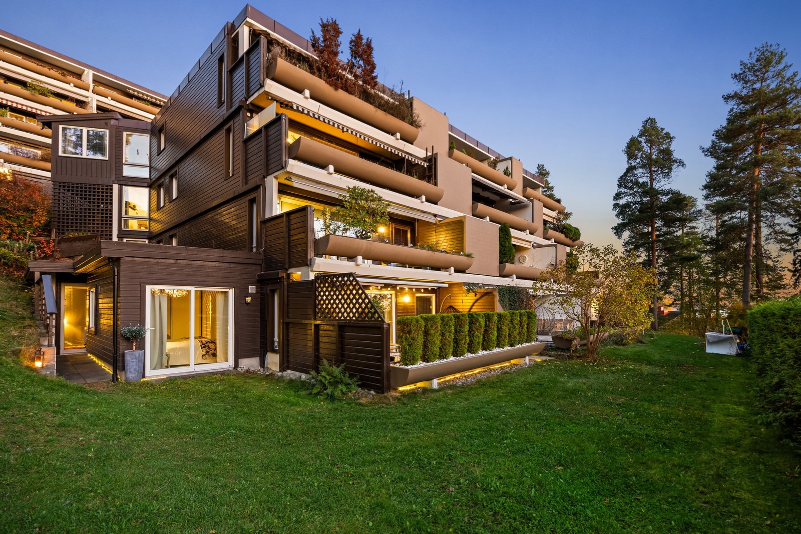 Sommerbilde (kveld) av Olav Aukrust vei 12. Det er direkte adkomst fra uteplassen til flott opparbeidet fellesareal. Egen, praktisk inngang fra bakkeplan til kjøkkenet Galleribilde