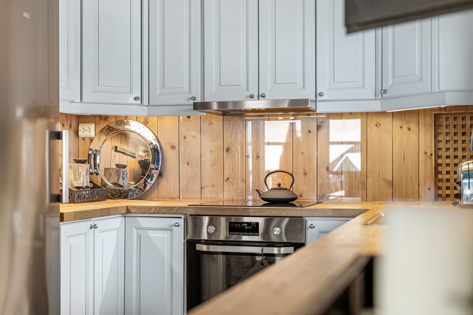 Lunt, delikat og praktisk. Et kjøkken som innbyr til bruk. Gjennomsiktig sprutsikring bak platetopp for ivareta den lune hyttestilen med treveggene. Galleribilde