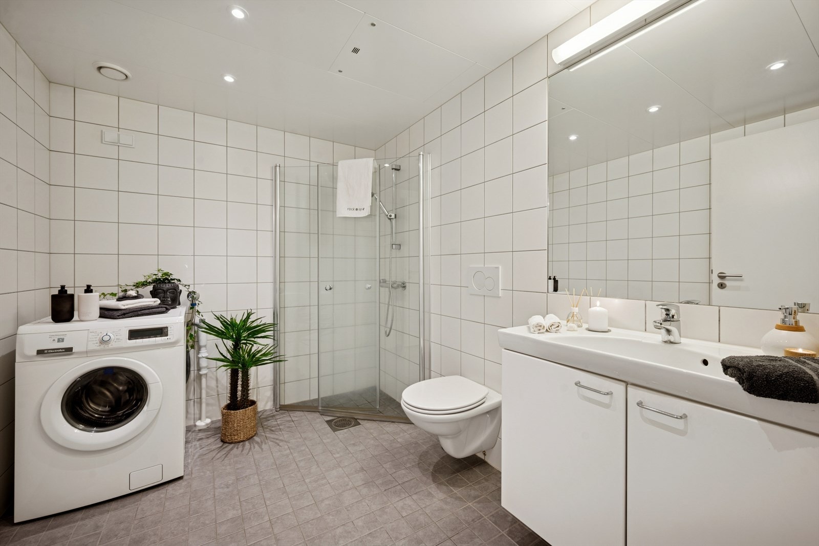 Badet har fliser på gulv og vegger. Skapinnredning, innfliset speil, veggfestet toalett, dusj med innfellbare vegger og opplegg for vaskemaskin. Galleribilde