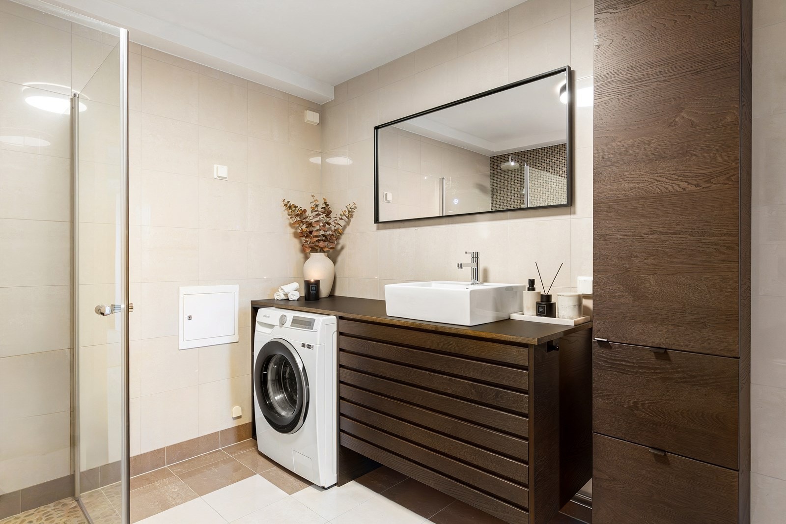 Badet fremstår moderne og delikat med store beige fliser på vegger og gulv som gir et rolig og helhetlig uttrykk. De varme flisetonene skaper en fin kontrast mot den mørke baderomsinnredningen, som tilfører rommet dybde og et eksklusivt preg. Galleribilde