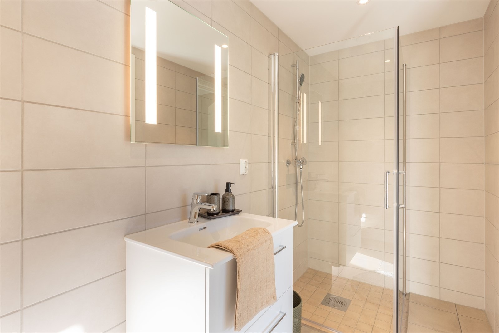 Lekkert helfliset bad som er innredet med baderomsinnredning med heldekkende servant, speil med belysning, vegghengt toalett og dusjhjørne med glassdører. Galleribilde