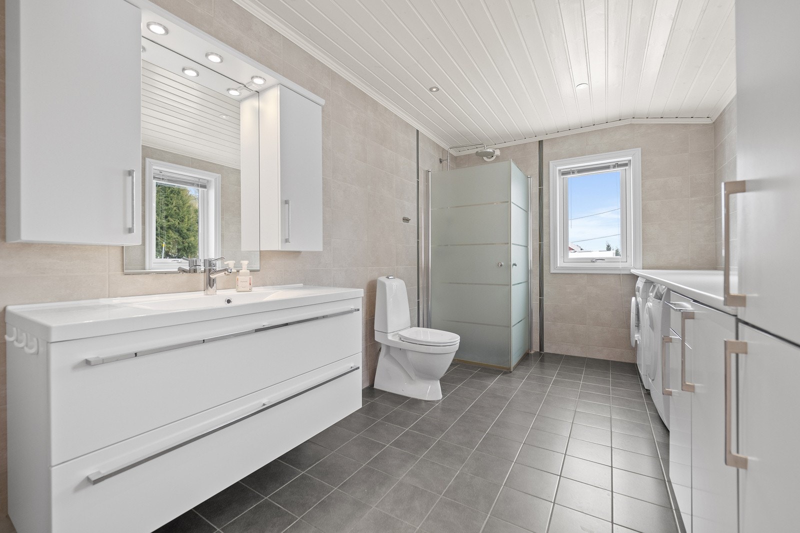 Flislagt bad med varme i gulv og panelbord i himling med downlights. Vegghengt servantinnredning med skuffer og ovenpåliggende servant. Galleribilde