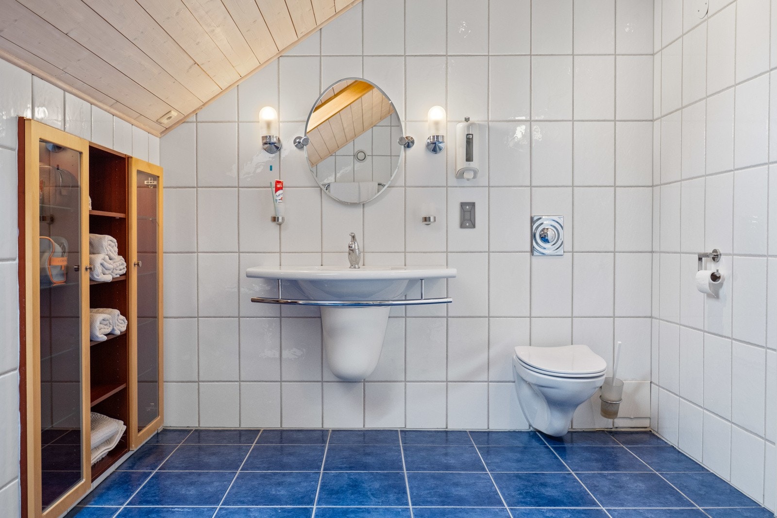 Badet er innredet med veggmontert servant og toalett, speil med belysning, og innebygde skap med hyller og glassdører. Galleribilde