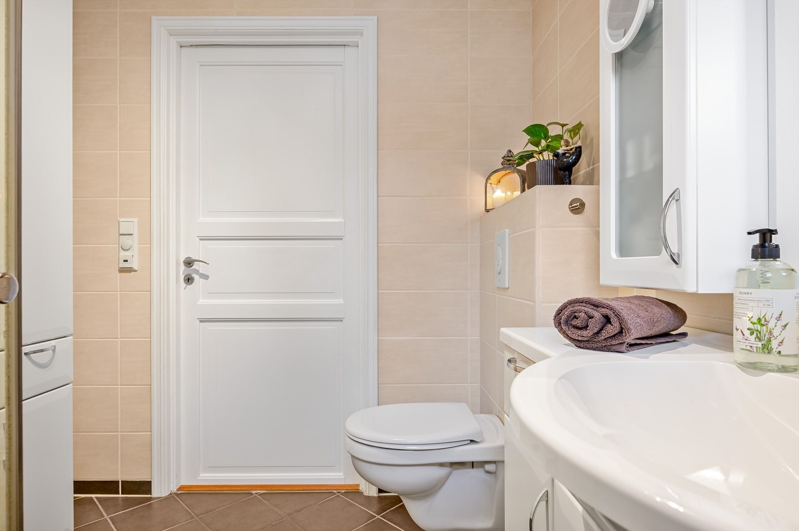 Lys baderomsinnredning med heldekkende servant, speil med belysning og vegghengte skap over. Baderommet er også utstyrt med ekstra høyskap, dusjhjørne med innfellbare glassdører og vegghengt toalett. Galleribilde