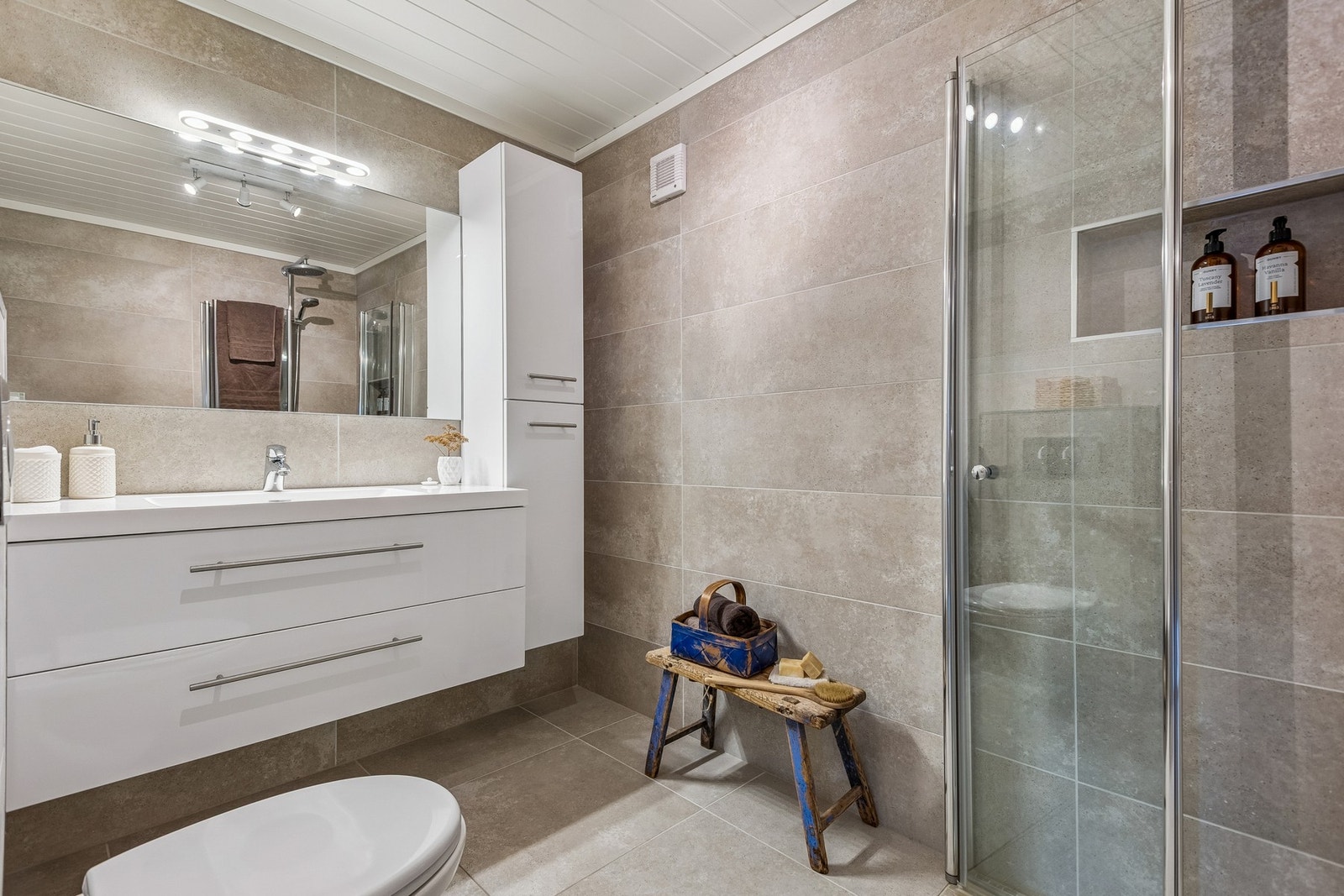 Badet er utstyrt med veggmointert toalett, servant med tilhørende skapinnredning og speil, innfellbare glassdører og integrert hylle i dusjsone. Varmekabler i gulv. Galleribilde