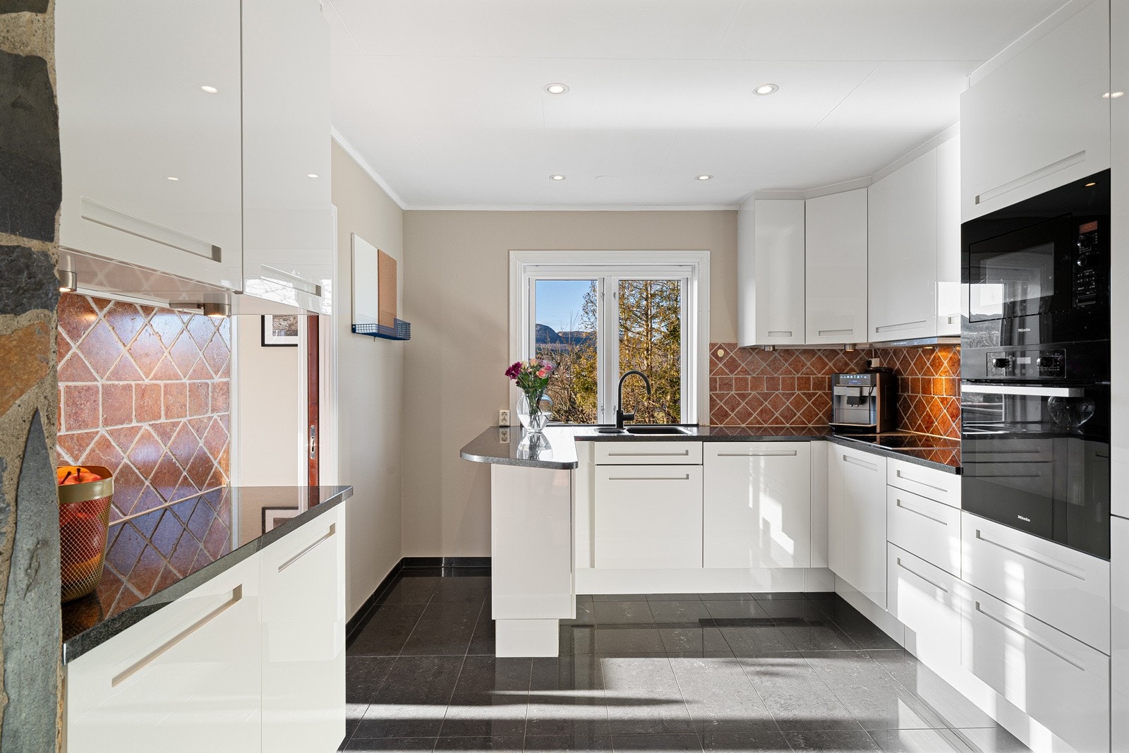 Innredningen har slette, hvite fronter som gir et tidløst og ryddig uttrykk, kombinert med en solid benkeplate i stein. Fliser mellom benkeplate og overskap fungerer som en praktisk og dekorativ detalj som binder helheten sammen Galleribilde