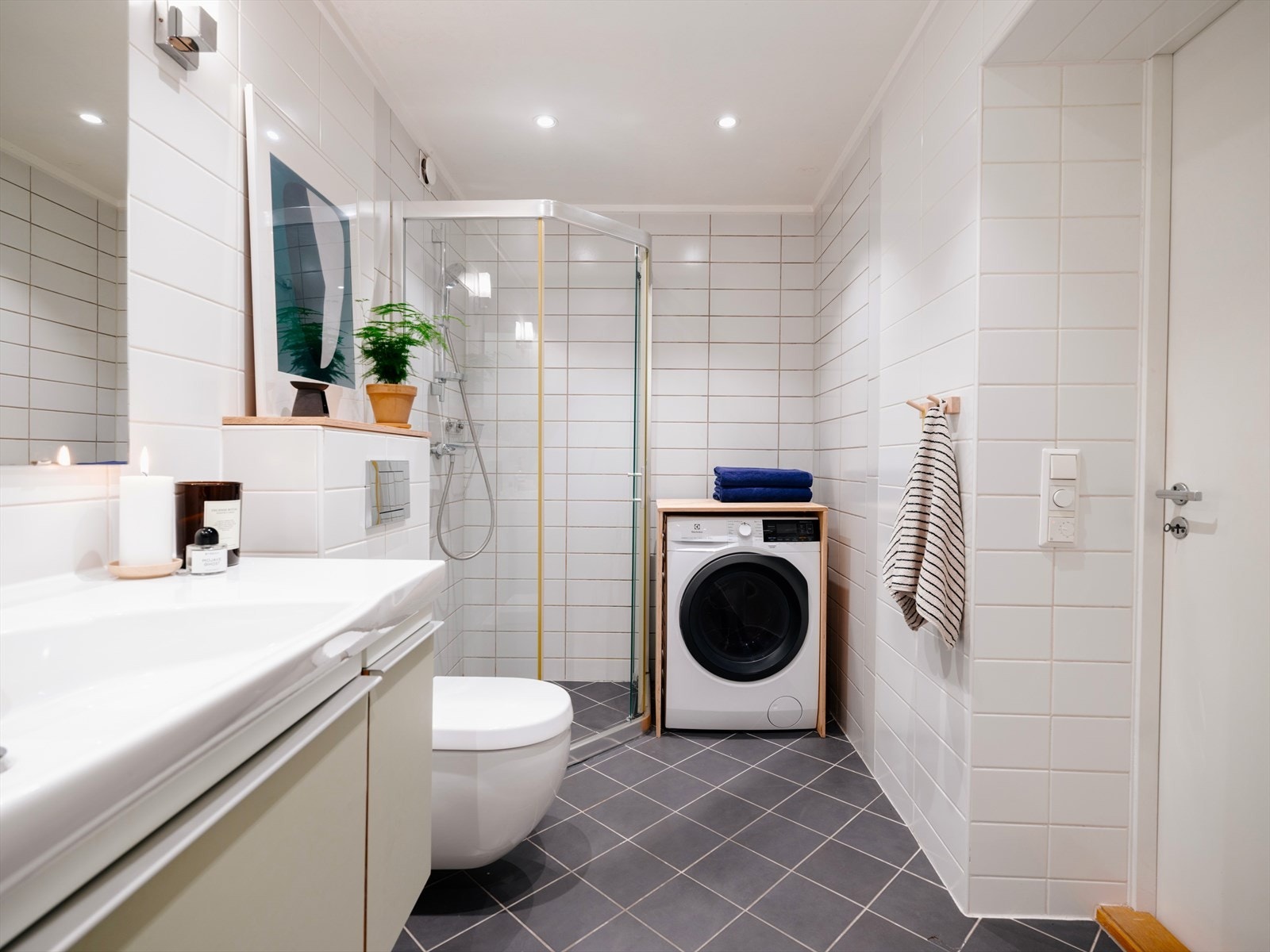 Pent flislagt bad av behagelig størrelse med dusjhjørne, klosett, servant med oppbevaring og opplegg til vaskemaskin. Galleribilde