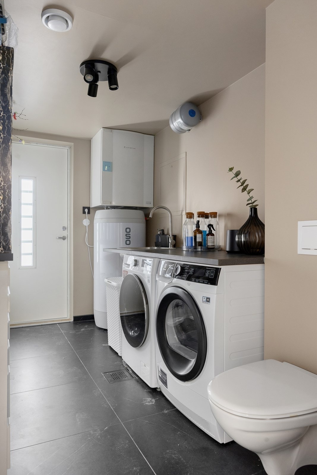 Vaskerom med fliser på gulv og lune fine farger på vegger og tak. Rommet fungerer også som teknisk rom/bi-inngang og er utstyrt med vegghengt toalett. Galleribilde