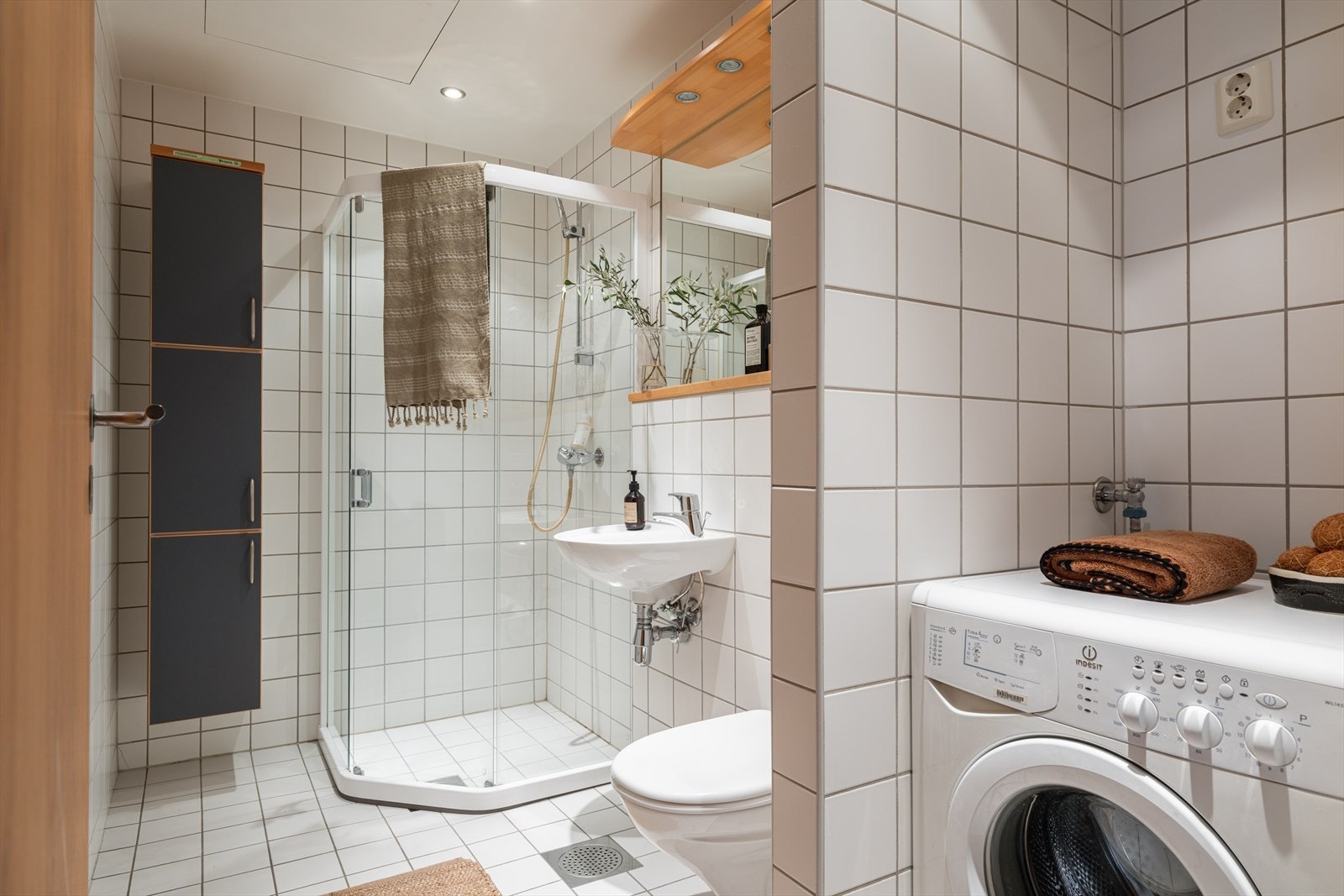 Bad/vaskerommet er flislagt med lyse fliser på gulv og vegger og har en praktisk planløsning med dusjsone og vaskesone. Galleribilde