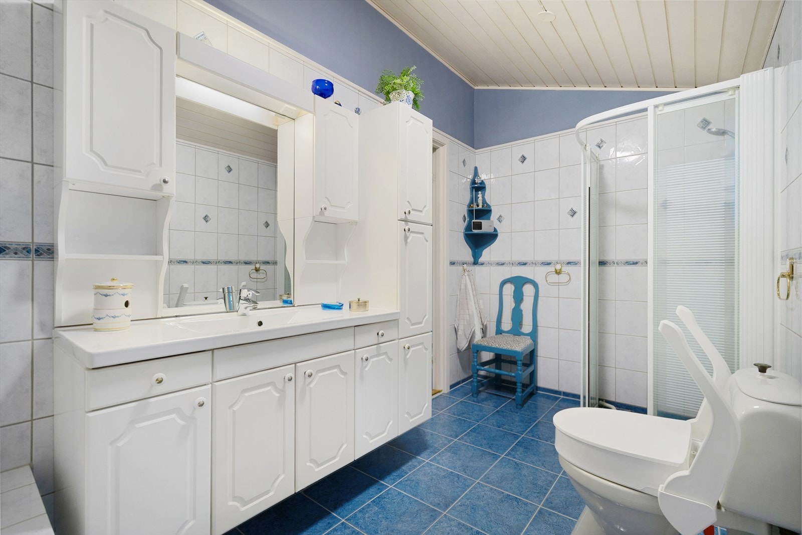 Flislagt bad med dusjhjørne, gulvmontert toalett og innredning med skap på begge sider av speil, et høyskap og porselensservant med benkeskap under og elektriske varmekabler i gulv. Galleribilde